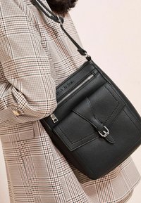 Borsa a tracolla nera in pelle con superficie testurizzata, chiusura con zipper e tracolla regolabile, abbinata a un cappotto a motivo check.