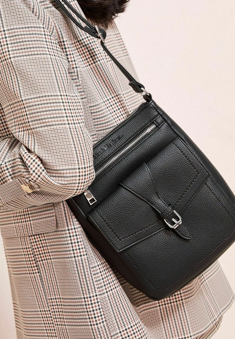Borsa a tracolla nera in pelle con superficie testurizzata, chiusura con zipper e tracolla regolabile, abbinata a un cappotto a motivo check.