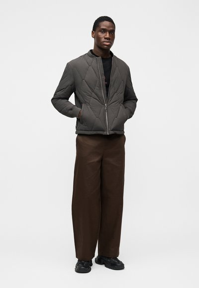 Chaqueta acolchada gris oscuro con cuello alto, cremallera frontal y bolsillos laterales, combinada con pantalones marrones de pierna ancha y zapatillas negras.