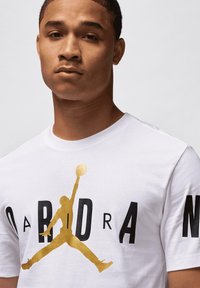 Jauns vīrietis valkā baltu T-kreklu ar lielu zelta Jumpman logotipu un melnu stilizētu "JORDAN" tekstu priekšā un uz piedurknes.
