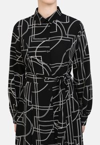 Black dress with white geometric patterns, long sleeves, a collared neck, and a belt at the waist. Features buttons down the front.