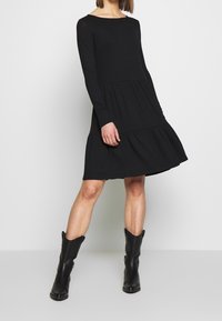 Svart långärmad klänning med ett åtsittande bodice och en flikad kjol, tillverkad av mjuk tyg. Buren med svarta ankelboots. Enkel, modern design.