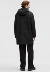 Mand, der står med ryggen til, iført en sort hættetrøje, sorte bukser med lommer og sorte sneakers mod en ensfarvet grå baggrund.