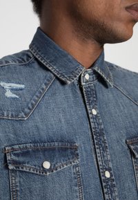 Farkkupaita keskiharmaassa sinisessä, jossa on jämäkkä kaulus, napitus ja kaksi rintataskua, joissa on painonapit. Olkapäällä kulutettu yksityiskohta.