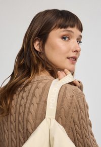 Maglione lavorato a maglia beige con motivo a trecce. Cinghia della borsa crema con testo "POLO CLUB". Modella con lunghi capelli castani, di schiena rispetto alla fotocamera.