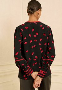 Blusa nera con stampa di labbra rosse, maniche a sbuffo e bordo ondulato rosso lungo il collo e i polsini. Tessuto liscio con leggero sheen.