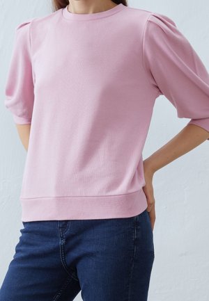 Vrouw die een lichtroze top met pofmouwen en donkerblauwe spijkerbroek draagt, staat met één hand op haar heup tegen een effen achtergrond.