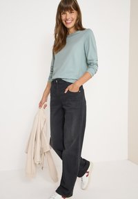Hellblau gestreiftes Langarmoberteil, kombiniert mit schwarzen, weit geschnittenen Jeans und weißen Sneakers mit roten Akzenten. Eine beige Jacke wird in der Hand gehalten.