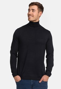Schwarzer gerippter Rollkragenpullover im taillierten Design, mit langen Ärmeln und einer weichen Textur. Über Jeans getragen, die Hände in den Taschen.