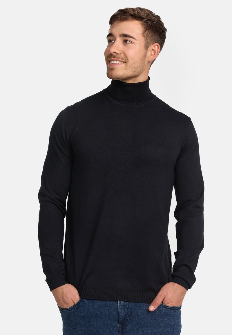Schwarzer gerippter Rollkragenpullover im taillierten Design, mit langen Ärmeln und einer weichen Textur. Über Jeans getragen, die Hände in den Taschen.