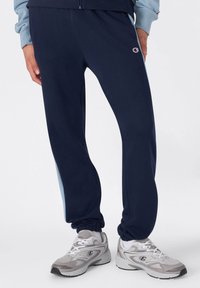 Pantalones de chándal azul marino con un corte entallado y puños elásticos, que presentan un panel lateral azul claro y un pequeño logo en la parte superior del muslo.
