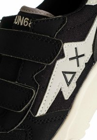 Sneaker nera con strappi in velcro, dettaglio a forma di fulmine bianco con triangoli neri e simboli più, e suola beige.
