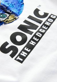 Bílá tričko s modro-zlatým flitrovým designem Sonicovy tváře, pod kterým je výrazný černý text "SONIC THE HEDGEHOG".