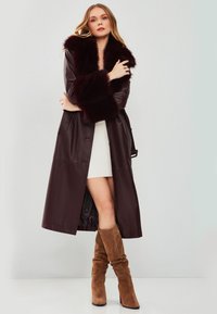 Cappotto lungo in pelle bordeaux con collo e polsini in pelliccia, caratterizzato da una texture liscia e due tasche frontali, indossato sopra un vestito bianco.