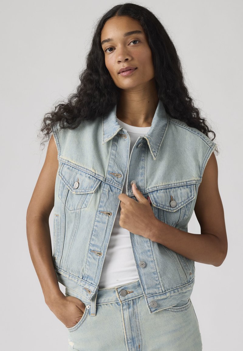 Femme aux longs cheveux bouclés portant un gilet en jean bleu clair sans manches sur un haut blanc, debout avec une main dans la poche.