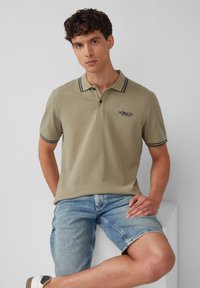 Oliwkowy polo z klasycznym kołnierzykiem, akcentami w granatowe paski oraz haftowanym logo. Połączony z jasnoniebieskimi szortami dżinsowymi.