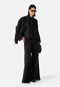 Nainen, jolla on musta helmillä koristeltu takki hartioilla, löysä musta paita, leveät mustat farkut, mustat aurinkolasit ja musta clutch-laukku.