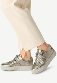 Sneaker metallici argento con una superficie liscia, punta rotonda, suola bianca piatta e chiusura con lacci, abbinati a pantaloni color crema.