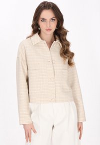 Beige ternetjakke med krave, lange ærmer, knaplukning og to frontlommer, parret med lyse bukser.