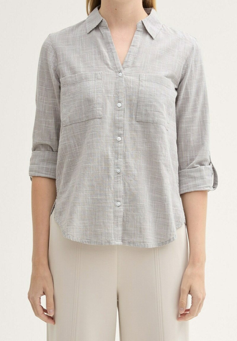 Femme portant une chemise à carreaux gris clair boutonnée avec des manches trois-quarts retroussées et un pantalon beige, debout devant un fond uni.