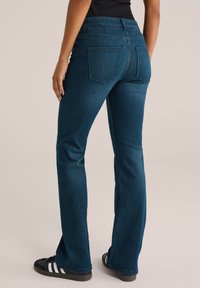 Temno modri flare jeans z gladko teksturo, z dvema zadnjima žepoma in pasom srednje višine. Nosi se z črnimi supergami.