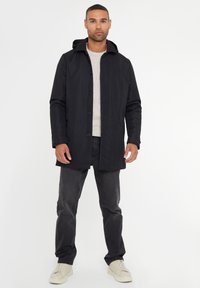 Zwarte waterdichte jas met een capuchon, met een rechte snit, knoopsluiting en een getextureerde binnenkant. G gedragen over een licht gebreid truitje en jeans.