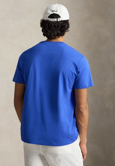 Άνδρας που φοράει βασιλικά μπλε t-shirt, λευκά σορτς και λευκό καπέλο με το κείμενο "POLO", στεκόμενος μπροστά σε ουδέτερο μπεζ φόντο.