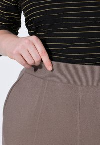 Jasnobrązowe spodnie z ribbowanym paskiem w talii, z fakturowanym wykończeniem, posiadające boczne kieszenie. Dłoń wskazuje na pas. W tle czarna bluzka w paski.