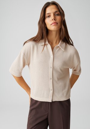 Junge Frau trägt ein beigefarbenes kurzärmeliges Hemd mit Knöpfen und dunkelbraune Hose, steht mit den Händen hinter dem Rücken vor einem schlichten grauen Hintergrund.