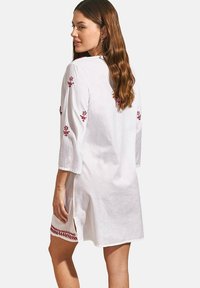 Robe en lin blanc avec des manches trois-quarts, ornée de broderies florales rouges sur les manches et l'ourlet, et avec des fentes sur les côtés pour plus de mouvement.