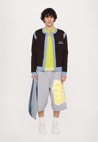 Black and blue reveal jacket with button closure, bright green shirt, gray shorts featuring yellow text, and white sneakers.