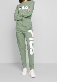 Hellgrünes Langarmoberteil und passende Jogginghose mit einem weißen Logoakzent. Das Material ist weich, hat einen lockeren Schnitt und einen elastischen Bund.