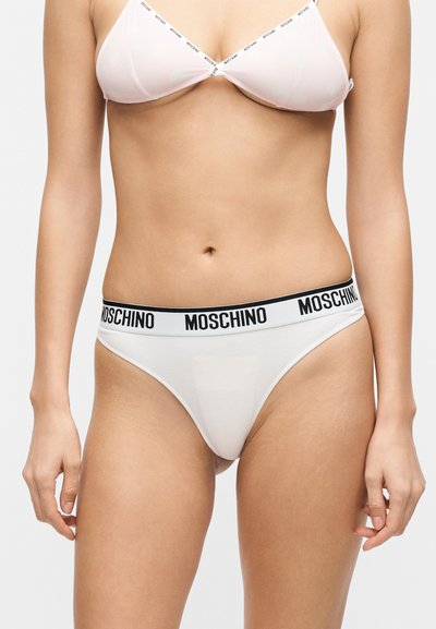 Γυναίκα που φοράει σετ λευκή εσώρουχα Moschino με μαύρο λογότυπο στις ελαστικές ταινίες, στέκεται μπροστά σε μονόχρωμο φόντο.