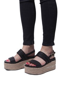 Sandalias de plataforma negras con suela de yute trenzada. Presentan dos correas anchas y una hebilla. Superficie texturizada, puntera abierta y esmalte rojo en las uñas.