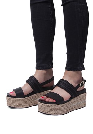 Sandalias de plataforma negras con suela de yute trenzada. Presentan dos correas anchas y una hebilla. Superficie texturizada, puntera abierta y esmalte rojo en las uñas.