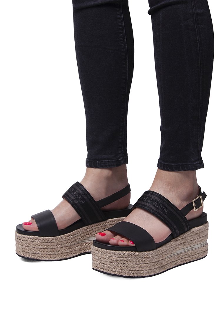 Sandalias de plataforma negras con suela de yute trenzada. Presentan dos correas anchas y una hebilla. Superficie texturizada, puntera abierta y esmalte rojo en las uñas.