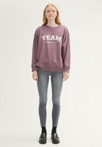 Lila kapucnis pulcsi, fehér "TEAM" felirattal, kerek nyakkivágással, hosszú ujjakkal, szürke, szűk farmernadrággal és fekete cipővel párosítva.