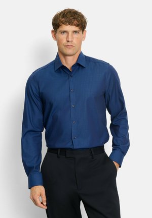 Mann mit kurzen, lockigen Haaren, der ein knöpfbares, dunkelblaues Langarmhemd trägt, das in schwarze Hosen gesteckt ist, und vor einem weißen Hintergrund steht.