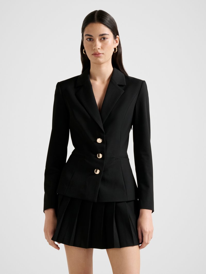 Μαύρο σακάκι με εφαρμοστή γραμμή, διακοσμημένο με τρία χρυσά κουμπιά, σε συνδυασμό με μαύρη πλισέ μίνι φούστα. Λείο ύφασμα και δομημένος σχεδιασμός.