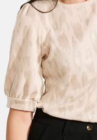 Beige lyhyt puff-hihainen pusero, jossa on hienovaraisia metallisia kuvioita, laitettuna mustien korkeavyötäröisten housujen sisään, joissa on nappikiinnitys.
