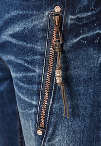 Jean en denim bleu foncé avec une fermeture éclair en cuivre ornée d'une tirette décorative, tissu texturé et légères altérations pour un détail supplémentaire.