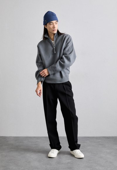 Gray half-zip sweater with ribbed cuffs, paired with black pants and white sneakers. Model wears a blue knit beanie. Simple, modern design.