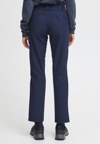 Pantalons de plein air bleu marine avec une texture lisse, une taille ajustée et des poches zippées, se rétrécissant légèrement à la cheville. Portés avec des chaussures noires.