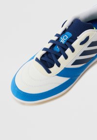 Zapatilla deportiva blanca y azul con una parte superior suave, lengüeta de malla texturada y cordones azules. Presenta tres rayas azules en el costado.
