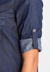 Camisa de denim azul oscuro con puños enrollados. Cuenta con un cierre de botón en el puño y un bolsillo en el pecho con un botón. Textura suave.