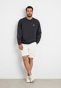 Tamsiai mėlynas džemperis su ilgomis rankovėmis, apykakle, ir mažai siuvinėta detalė ant krūtinės, derinamas su baltais šortais ir baltais sportbačiais.