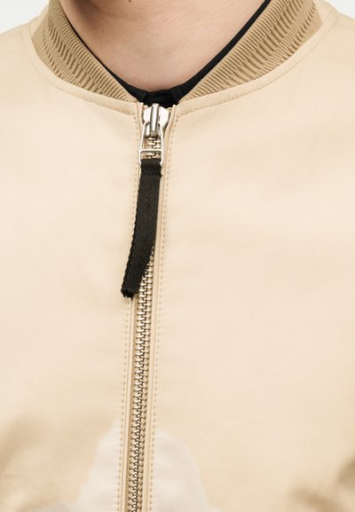 Közelkép egy bézs zakó cipzárjáról fekete szövet húzófüllel, bordázott gallérral, valamint a nyak és a fekete belső ing részével látható.