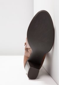 Botte de robe en cuir marron avec semelle noire texturée et talon bloc, inclinée contre un fond d'angle blanc.