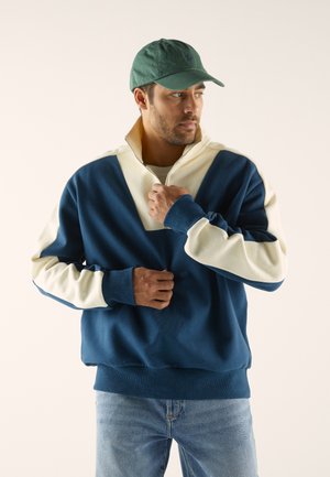 Pull dépasse en bleu marine et crème avec un col montant, fermeture éclair et poignets côtelés. Porté avec un jean bleu clair et une casquette verte.