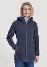 Vrouw in een donkergrijze hoodie met ritssluiting en lichtpaarse coltrui, met één hand in de zak van de jas, staand tegen een effen achtergrond.